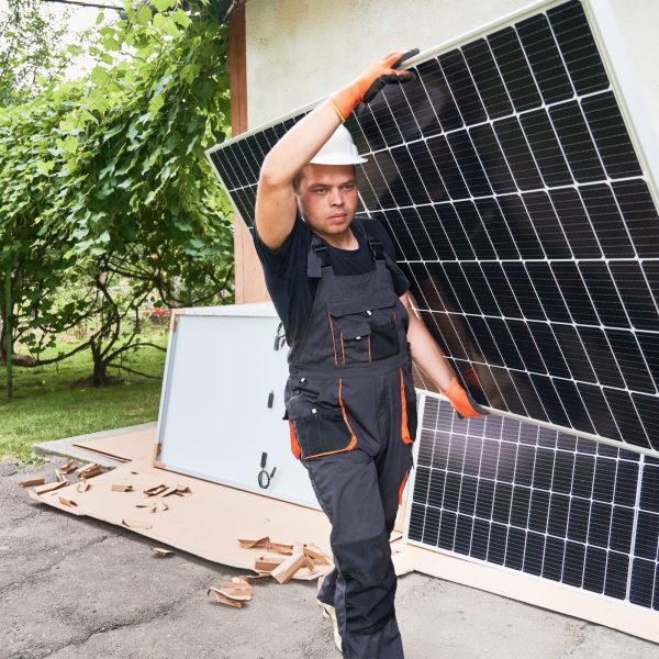 Serious man solar installer carrying solar module in the yard. Male worker in work overalls holding photovoltaic solar panel and walking down asphalt pathway outside the house.